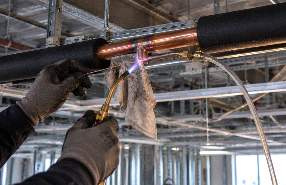 Technician brazing a refrigerant branch joint on insulated copper piping above a ceiling grid in an unfinished commercial interior